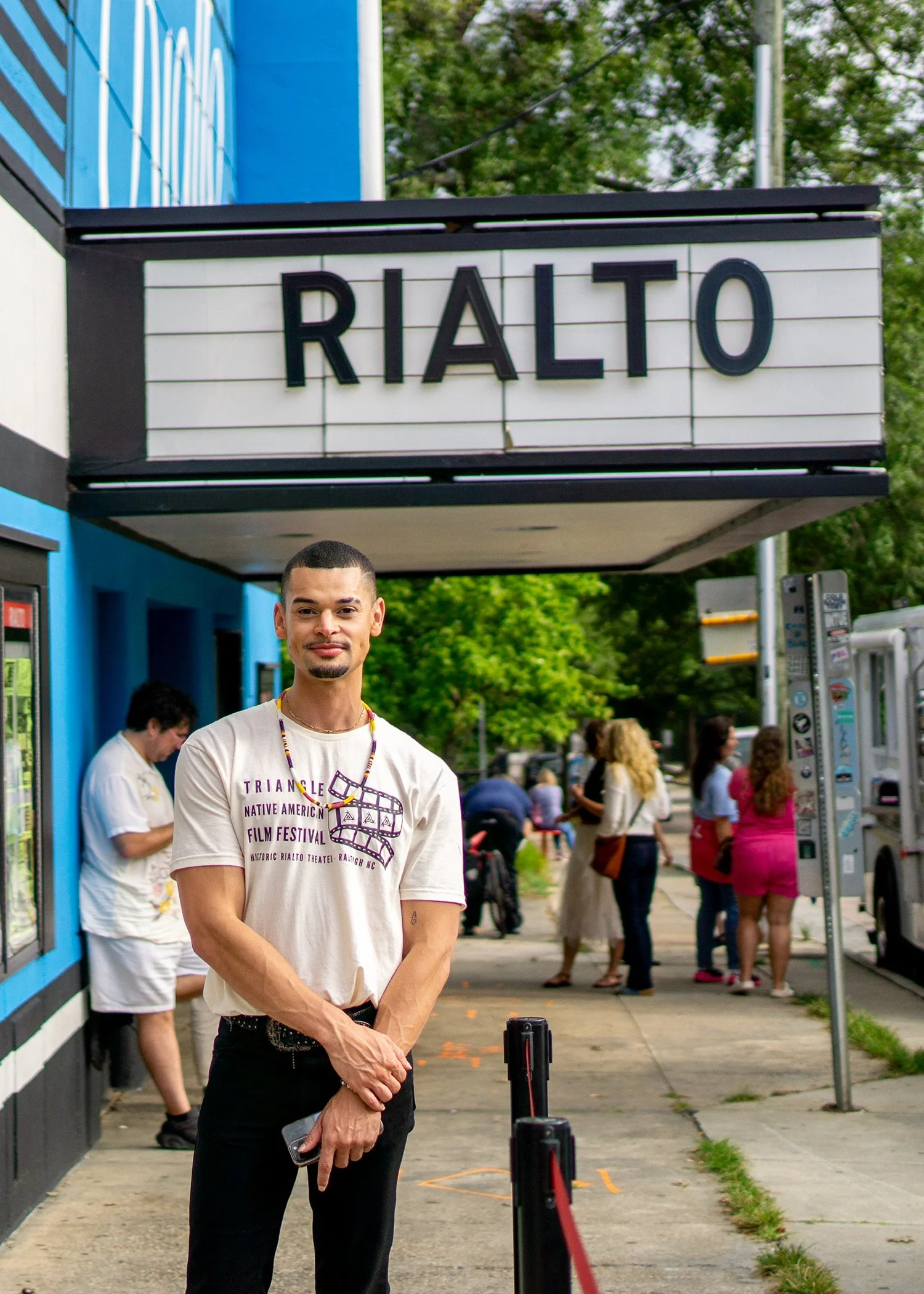 A man wearing a Triangle Native American Film Festival T-shirt stands outside the Rialto Theatre in Raleigh as attendees gather on the sidewalk behind him